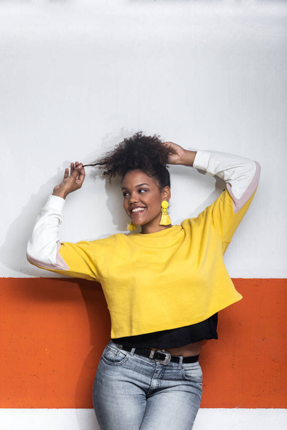 Positive African American millennial female in trendy yellow shirt touching curly hair and looking away while dancing near wall - Foto, imagen
