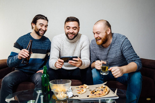 Group of three friends drinking beer and watching funny video on smartphone and sitting on the sofa at home, talking laughing and enjoying their time using smartphone - Zdjęcie, obraz