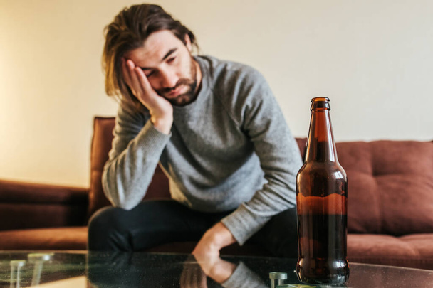 Young man suffering from strong headache or migraine sitting on sofa with bottle, millennial guy feeling intoxication and pain touching aching head, morning after hangover concept - Photo, image