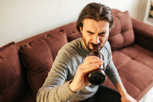 top view of young depressed drunk man drinking beer from the bottle, at home sitting on bed thoughtful and confused as alcoholic suffering alcoholism problem and alcohol addiction - Zdjęcie, obraz