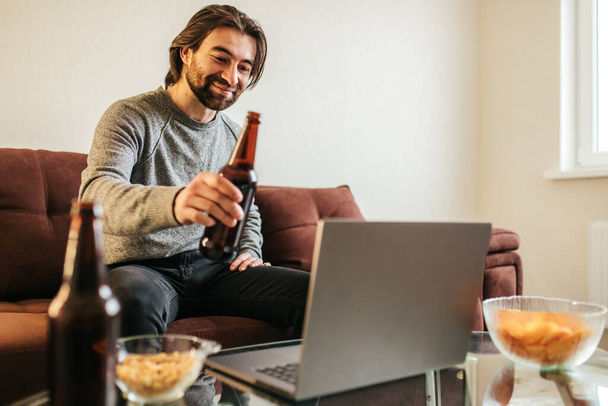 Young attractive caucasian man celebrating birthday online with bottle of beer. male sitting by laptop communicating videochat conference smiling cheerfully on webcam. Holiday alone. Quarantine. Social distance - Foto, imagen