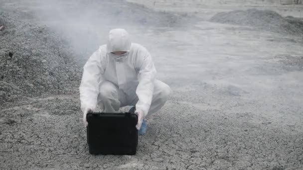 Un técnico de laboratorio con una máscara y un traje de protección química abre una caja de herramientas en tierra firme, alrededor del humo tóxico - Imágenes, Vídeo