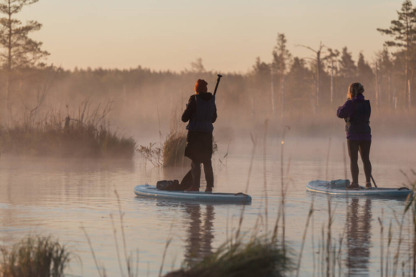 Grupo de jóvenes disfrutan haciendo SUP stand up paddle boarding al amanecer en el lago. Actividad temprano en verano - Foto, Imagen