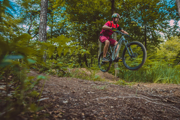 Side view of young caucasian male jumping with a mountain bike over a jump on a singletrail. Epic ride with a modern mountain bike on a trail park. - Photo, Image