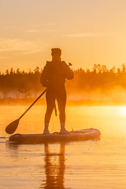 Silhouette de la jeune femme asiatique faisant SUP stand up paddle boarding au lever du soleil dans le lac. Activité matinale tôt en été - Photo, image