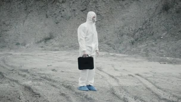 Lab technicus in een masker en chemisch beschermend pak, loopt op droge grond met een gereedschapskist door giftige rook - Video
