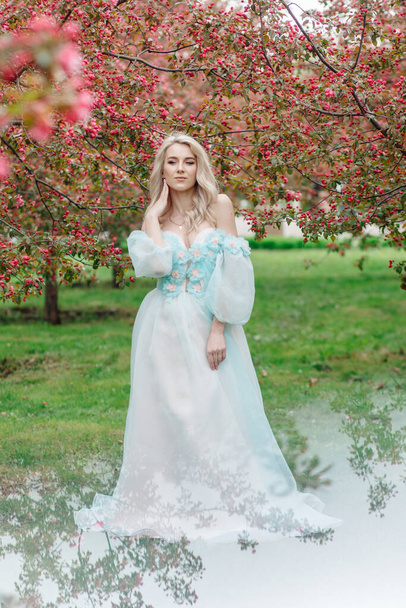 A blonde woman in spring in a pink blooming garden and in a beautiful blue dress. At the bottom of the photo is a mirror image - Photo, Image