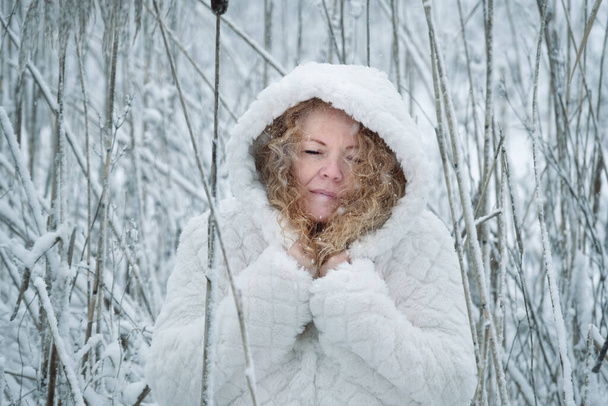 白いフードのコートを着た赤髪の熟女は雪の葦の冬の寒さの中で満面の笑みを浮かべて立って - 写真・画像