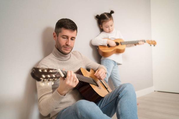 Папа играет на гитаре со своей дочерью. Ребенок учится играть на музыкальном инструменте с репетитором. Музыкальный дуэт отца с ребенком - Фото, изображение