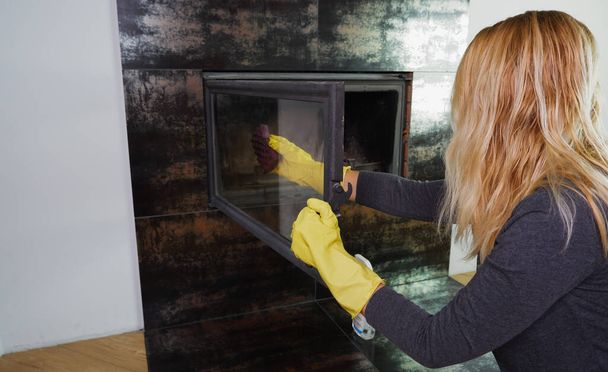 Sarı eldivenli bir kadın şöminenin camını yıkıyor. Günlük ödev günlük kış rutini - Fotoğraf, Görsel