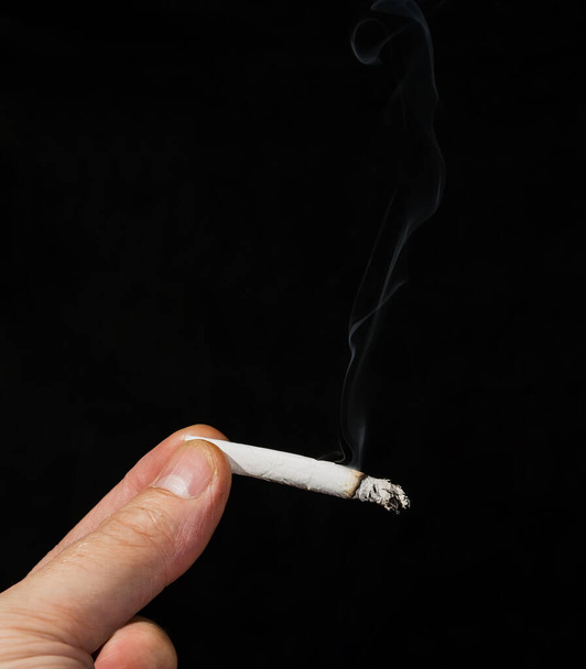 Hand holding a marijuana cigarette in front of a black background.The joint is lit, and smoke rises from it. There is room for text at the top. - Photo, Image