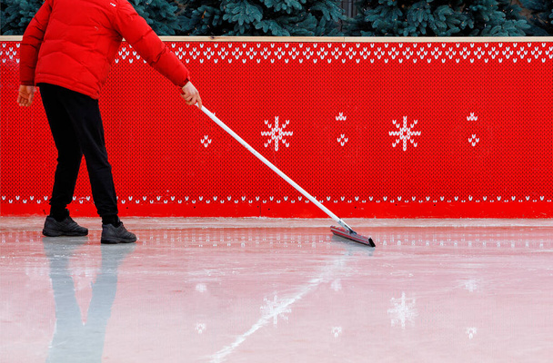 Работник ледяного стадиона, окруженный красным забором и одетый в красную куртку, очищает поверхность льда прорезиненной шваброй, чтобы удалить мелкие щепки. Копирование пространства. - Фото, изображение