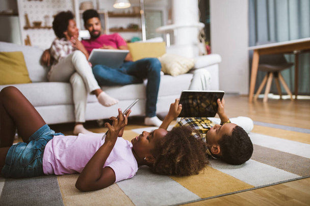 Menino e menina afro-americanos deitados no chão e usando tablet digital. Família em casa. - Foto, Imagem