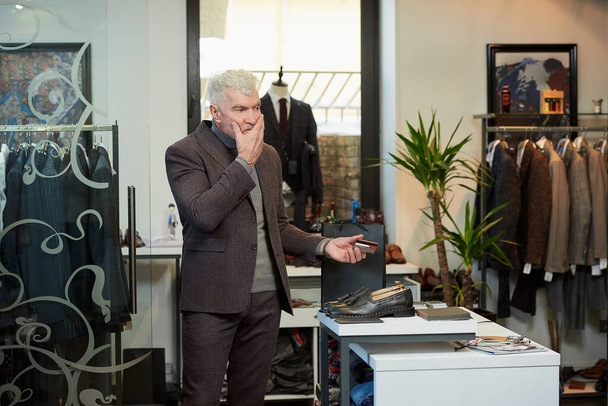 A mature man with gray hair and a sporty physique is holding a credit card to pay for purchases in a clothing store. A customer with a beard is massaging his face in a boutique - Photo, Image