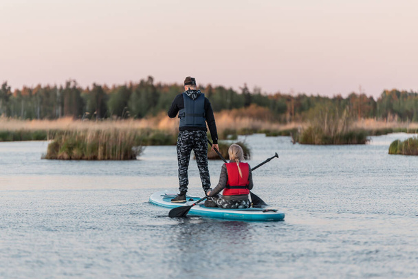 Pareja en SUP de pie paddle boarding al atardecer en el lago o pantano. Actividad vespertina - Foto, Imagen