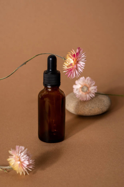 Brown glass bottle dropper with cosmetic oil with pink dry flowers on terracotta background front view, closeup - 写真・画像
