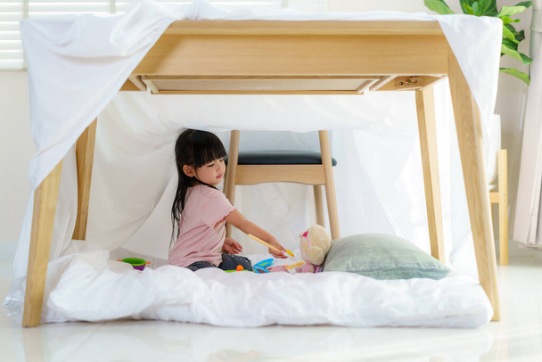 Asian cute little girl play cooking and feeding food to her doll while sitting in a blanket fort in living room at home for perfect hideout away from their other family members and for them to play imaginatively. - Foto, Imagem