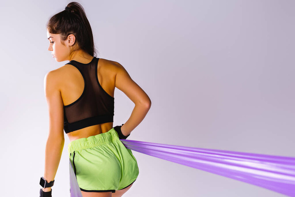 Photo from behind. A girl in fitness shorts and a top trains body mice with an resistance elastic band holding on to a white background with empty space for advertising. - Φωτογραφία, εικόνα