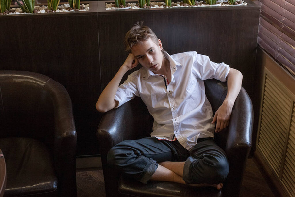 A boy in a white shirt and jeans sits in a chair. Teenager resting in a cafe - Foto, immagini