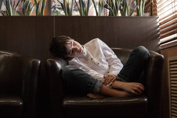 Chlapec v bílé košili a džínách sedí v křesle. Teenager odpočívá v kavárně - Fotografie, Obrázek