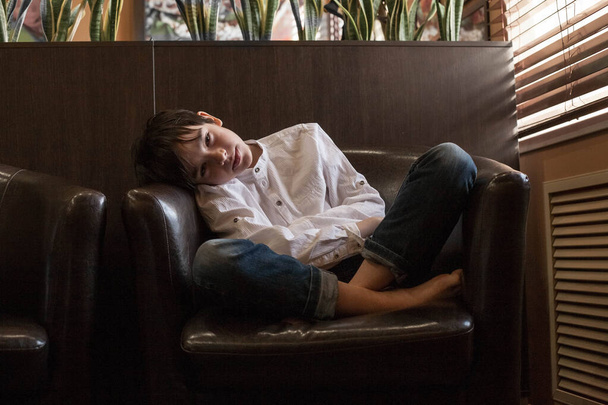 A boy in a white shirt and jeans sits in a chair. Teenager resting in a cafe - Fotografie, Obrázek