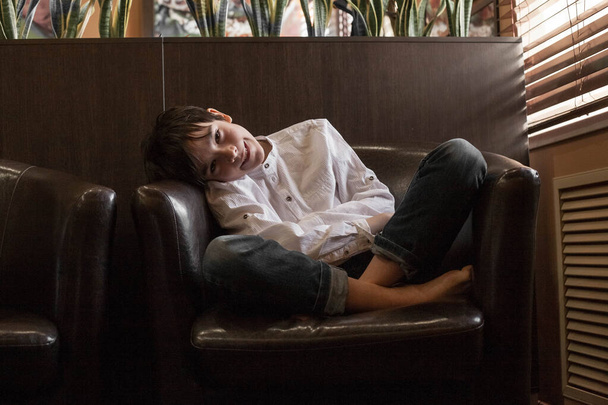 A boy in a white shirt and jeans sits in a chair. Teenager resting in a cafe - Φωτογραφία, εικόνα