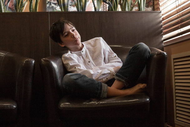 A boy in a white shirt and jeans sits in a chair. Teenager resting in a cafe - Фото, изображение