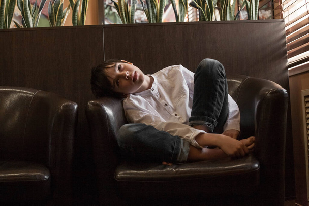 A boy in a white shirt and jeans sits in a chair. Teenager resting in a cafe - 写真・画像