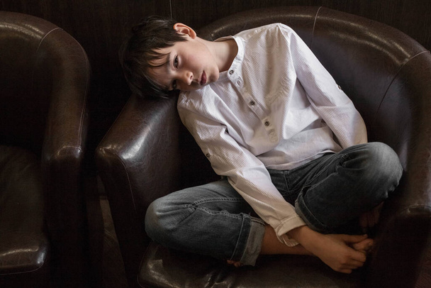 A boy in a white shirt and jeans sits in a chair. Teenager resting in a cafe - Фото, зображення
