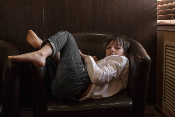 A boy in a white shirt and jeans sits in a chair. Teenager resting in a cafe - Zdjęcie, obraz
