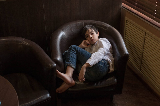 A boy in a white shirt and jeans sits in a chair. Teenager resting in a cafe - Foto, Bild