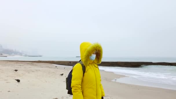 Aikuinen nainen lämpimässä keltaisessa takissa, jossa on reppu, käyttää suojaavaa lääketieteellistä naamiota, kävelee pitkin merenrantaa ja nauttii kauniista merinäköalasta. Kokonaiskuva COVID-19-pandemiasta - Materiaali, video
