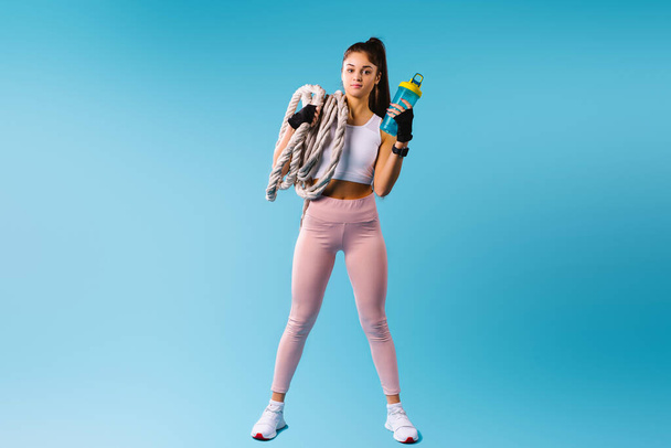A muscular fitness girl in sport tight leggings holds a rope knot and a protein shake on her shoulder. Posing against a blue background with empty space. - Foto, immagini