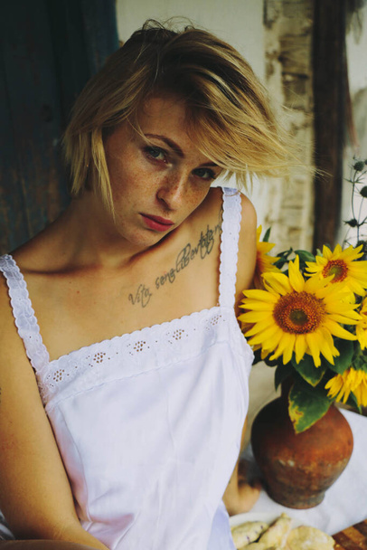 Portrait of a funny sexy blonde woman with blond hair eating at a table on the porch of an old village house. A bouquet of sunflowers. - 写真・画像