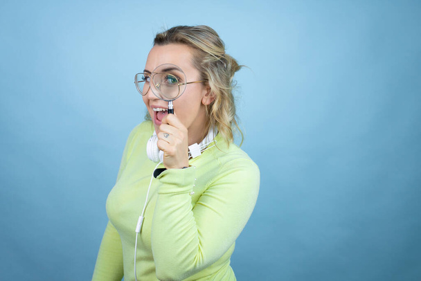 jonge blanke vrouw het dragen van een koptelefoon op de hals over blauwe achtergrond verbaasd kijken door een vergrootglas - Foto, afbeelding