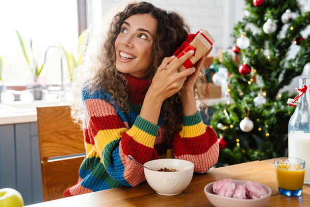 Joyful charming woman holding gift box while having breakfast in cozy kitchen - 写真・画像
