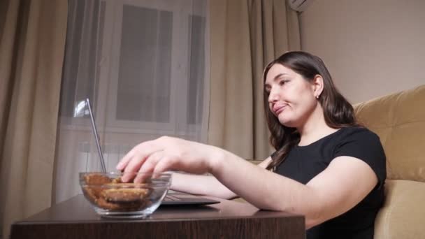Brunette in black t-shirt eats cookies and types on laptop - Footage, Video