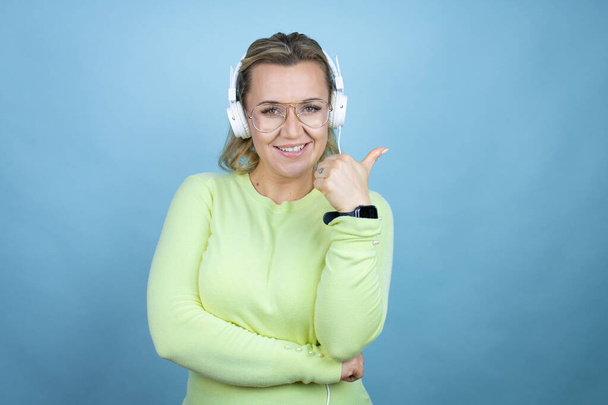 Giovane donna caucasica ascoltare musica utilizzando cuffie su sfondo blu sorridente e puntando con mano e pollice a lato - Foto, immagini