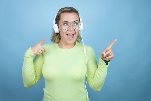 Giovane donna caucasica ascoltare musica utilizzando cuffie su sfondo blu sorpreso e puntando le dita lato - Foto, immagini