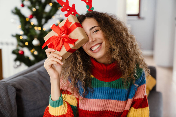 Joyful charming woman in toy deer horns holding gift box while sitting on couch at cozy room - Photo, Image