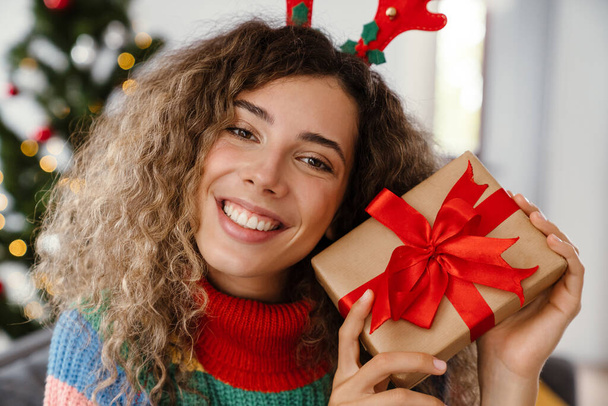 Joyful charming woman in toy deer horns posing with gift box at cozy room - Foto, Imagen