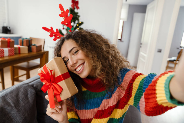 Joyful charming woman smiling and taking selfie photo with gift box at cozy room - Фото, изображение
