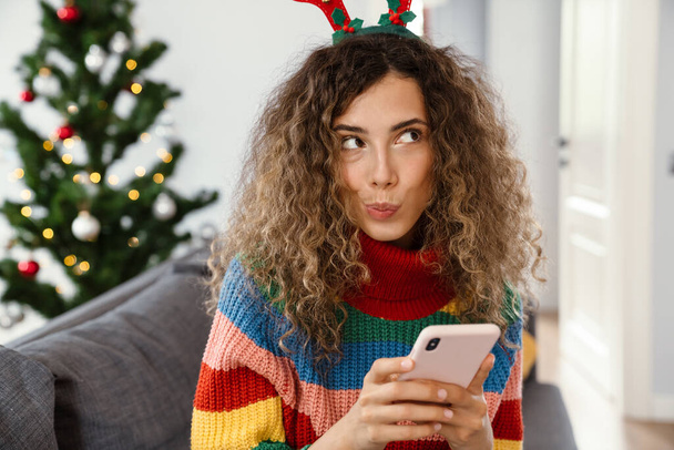 Thinking charming woman in toy deer horns using cellphone while sitting on couch in cozy room - 写真・画像