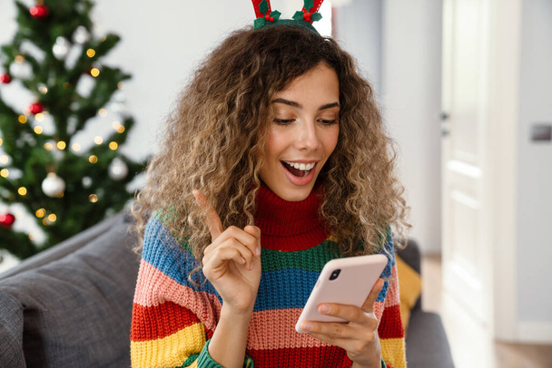 Joyful charming woman in toy deer horns using cellphone while sitting on couch in cozy room - Photo, image