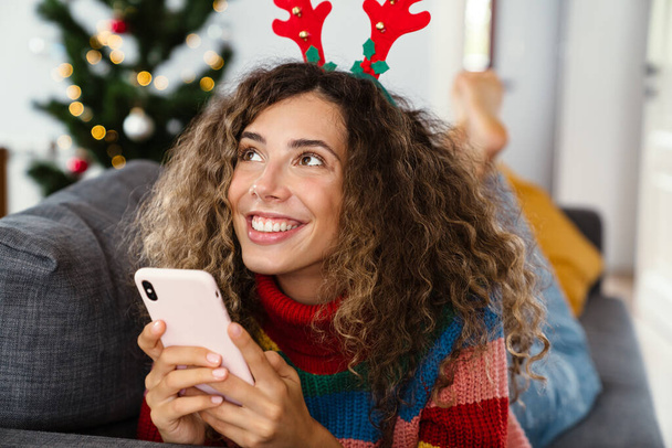 Happy charming woman in toy deer horns using cellphone while lying on couch in cozy room - Фото, изображение