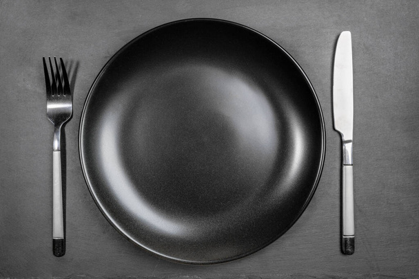 Empty blank black plate with fork and knife on black background of shale serving board. Mock up. Top view. - Photo, Image