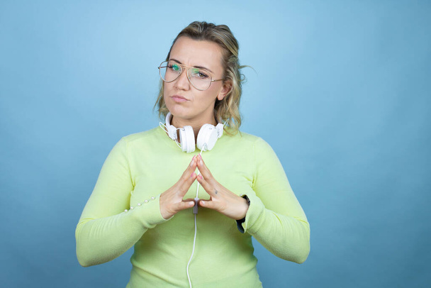 Mulher branca jovem usando fones de ouvido no pescoço sobre fundo azul com as mãos juntas e dedos cruzados sorrindo relaxado e alegre. Sucesso e otimista - Foto, Imagem