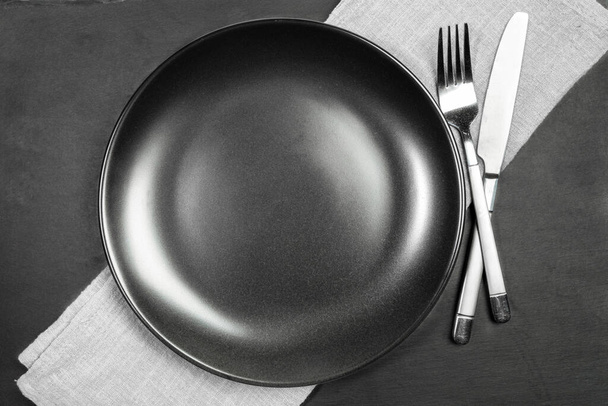 Empty blank black plate and cutlery on grey diagonal cooking towel on black shale serving board. - Foto, Imagen