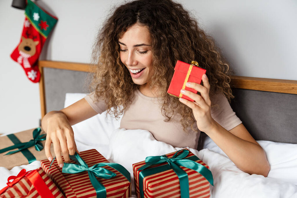 Feliz mujer agradable sonriendo y abriendo cajas de regalo mientras está sentado en la cama en casa - Foto, Imagen