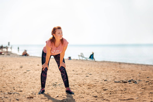 Eine erwachsene, müde schöne Frau in Sportbekleidung macht während eines Trainings eine Pause. Im Hintergrund das Meer und der Strand. Kopierraum. Draußen. - Foto, Bild